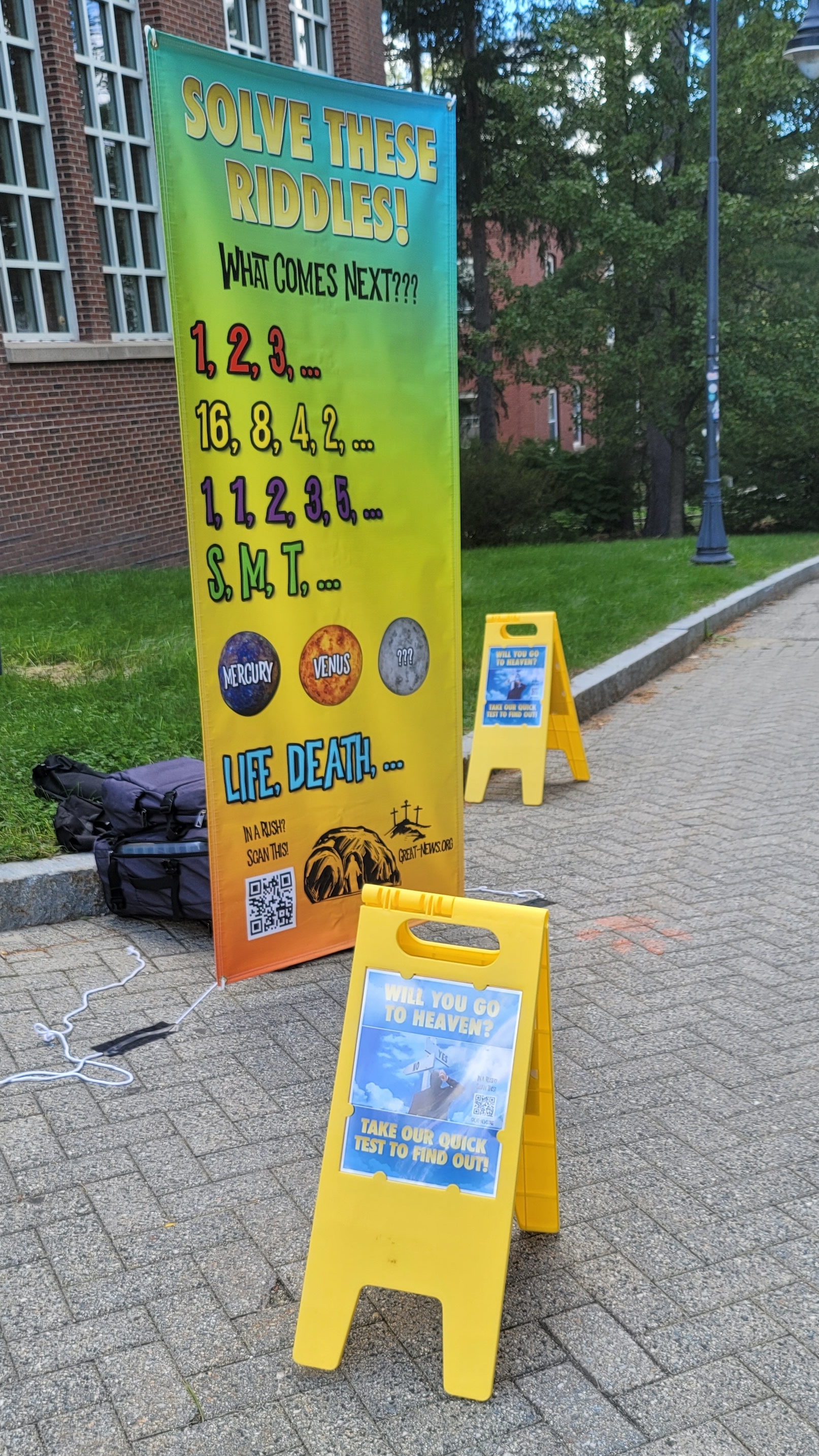 This is how we setup on campus. Many students stop to check out the large banner with the riddles. And the smaller A-Frame signs are helpful too.  All of them have "QR" codes on them, so if a student scans it with their phone, it will take them to our evangelistic website, https://great-news.org.   It's not at all uncommon for us to see students scanning the QR code.